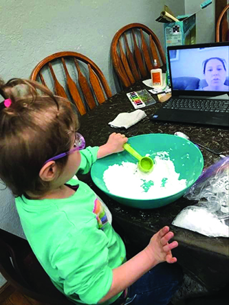 A White preschool student wearing purple glasses mixes flour in a green bowl as the TVI watches on Zoom. 
