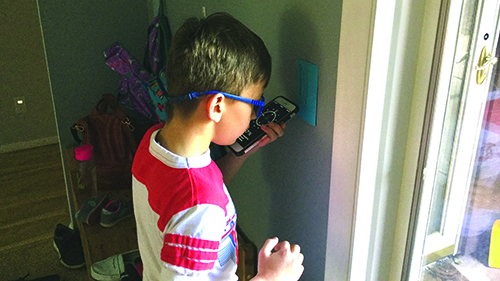 A White elementary school-aged boy wearing blue glasses holds an iPhone with a compass visible. The compass is pointing north and a blue card on the wall is visible with the letter "N" printed on it. 