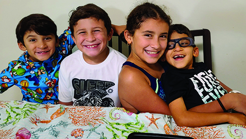 Three boys and a girl who are multiracial are celebrating a birthday. One boy sits on his sister's lap. He wears glasses and is missing his front teeth. All four siblings are smiling. 