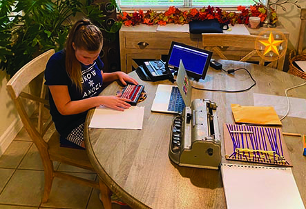 A White teenage girl is at her kitchen table with an abacus during online math instruction. A braille notetaker with an external monitor, a laptop, a Perkins brailler along with art materials sit on the table. 