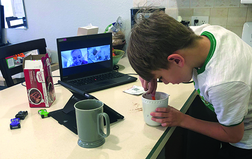 A White elementary student mixes ingredients in a mug to make a cake. His White female O&M specialist is giving him directions via Zoom. 