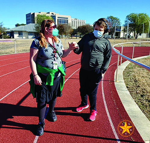 A White teenager and a White teaching assistant are on a track. He is wearing sound muffling headphones. The teaching assistant is guiding the teenager during the school’s Jingle Bell Run. She has 2 jingle bells around her neck to signify they have completed 2 laps around the track. 