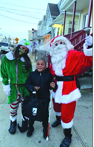 A Black elementary school student poses outside with Santa and an elf. The student has a cane, Santa wears sunglasses and the elf wears a mask. 