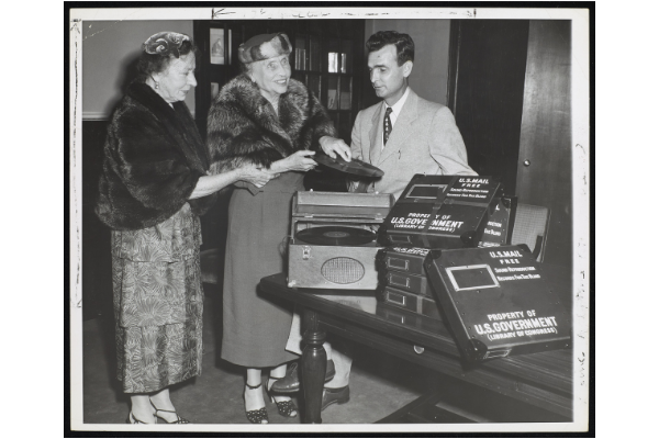 Polly Thomson and Helen Keller with M. Robert Barnett at AFB. Keller examines a Talking Book. A Talking Book machine is beside her on the table, as are many of the U.S. Government boxes that the records were shipped in. Thomson and Keller are wearing ankle length dresses, hats and fur coats.