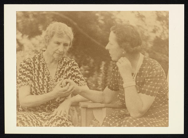 Candid close-up photograph of Helen Keller and Polly Thomson sitting outdoors in wooden chairs. They both use their right hands to tactile fingerspell with each other and they are leaning towards each other as they communicate. Keller's expression reflects concentration. Thomson, whose head is turned to face Keller has her left arm raised against her chin. They both wear patterned dresses. Trees are visible in the background.