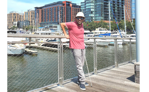 Amir Rahimi standing on the pier of Little Island. Boats are docked in the waters behind him. In the background, a row of buildings are capped by a clear, blue sky.
