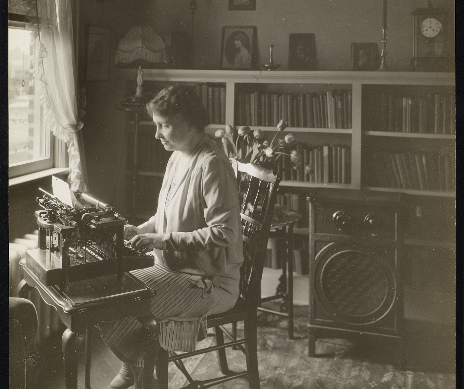 Helen Keller sits in a wooden chair at a small writing desk. She is using a typewriter that sits atop the desk. She is lit by sunlight shining through a window at her right. Behind her is a row of shelves filled with books. In front of the shelves is a floor-model radio. 