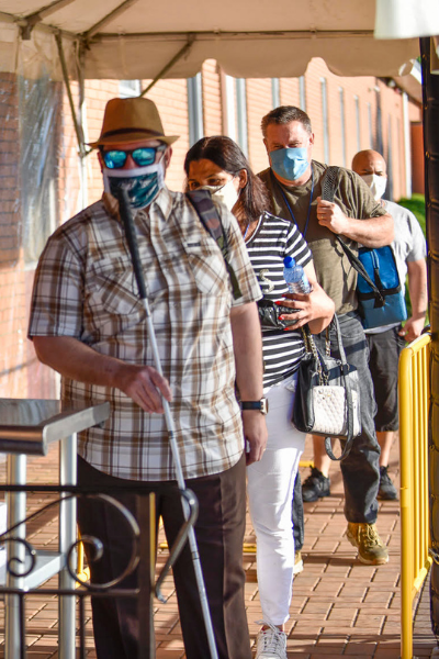 People standing in a line on the sidewalk out front of a brick building, waiting to enter. They all wear masks. The man at the front of the line holds a white cane used for navigation.