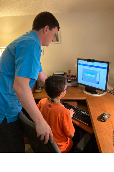 A bespectacled young boy sits at a desk situated in a corner of a room in a house. Standing behind him, looking over his shoulder, is his father. They are working together on a project on a computer. 