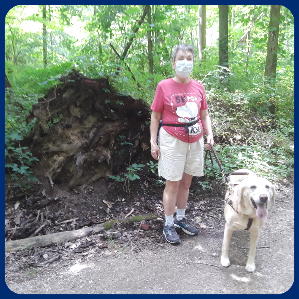 A woman and her service dog stand on a dirt path next to an uprooted tree in the woods.