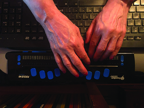A view from above of the hands of a person with light skin reaching over a laptop keyboard to rest on a braille display