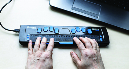 Fair skinned hands poised on a braille display that is connected to a laptop computer