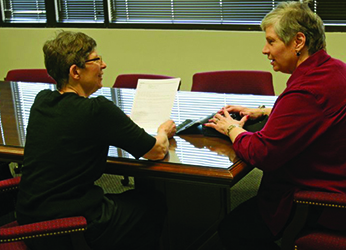 Two women with light skin and graying hair sit at a conference table, one woman using a braille notetaker as they chat