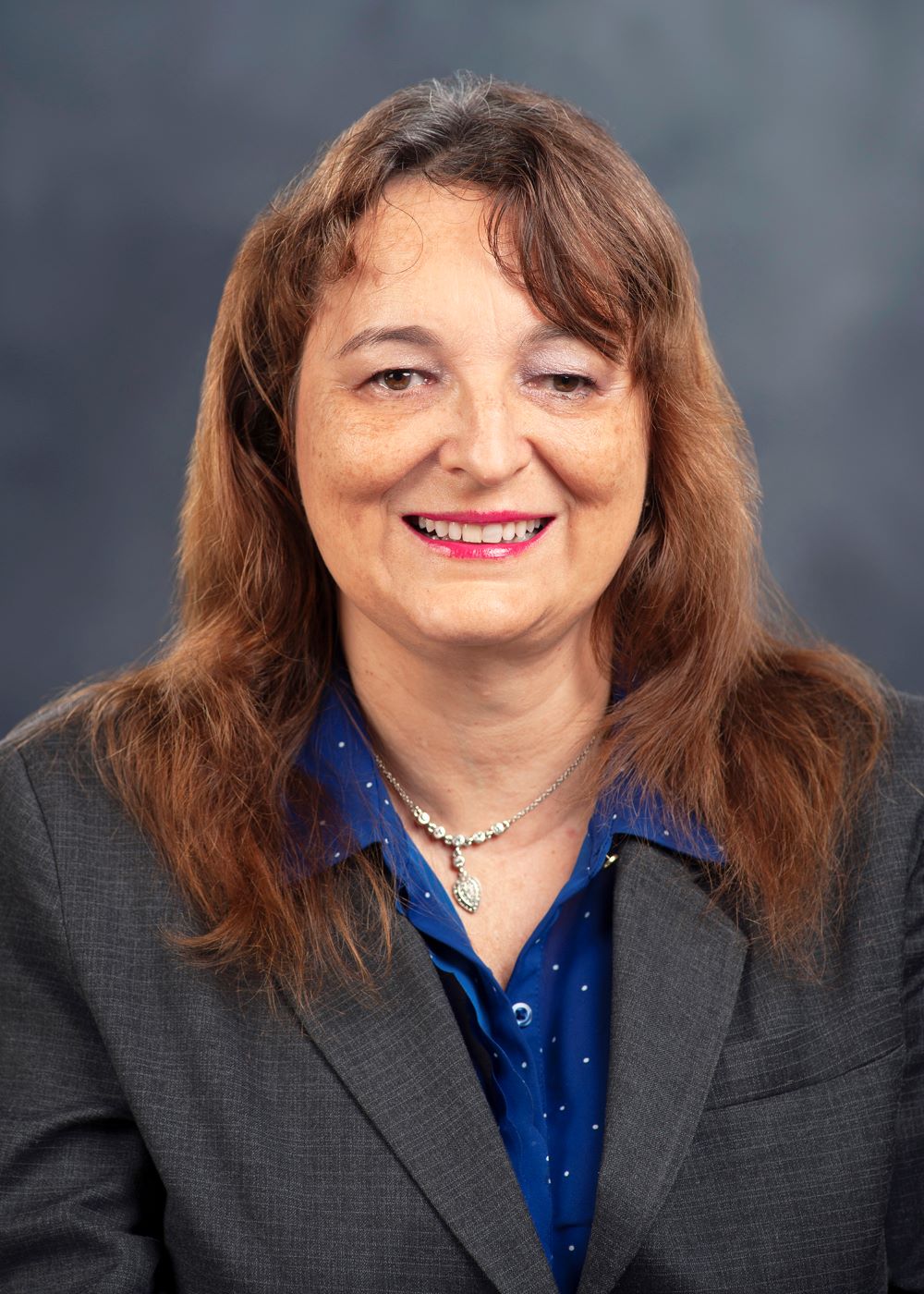 Headshot of Sylvia. Long brown hair, wearing a gray suit with blue blouse.