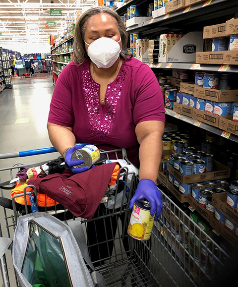 A woman is pictured at a grocery store. She is wearing a mask and blue gloves while placing cans into her shopping cart.