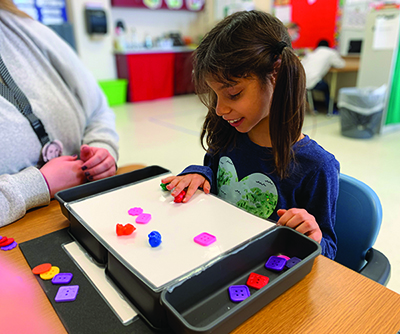 An elementary girl with ponytails placing magnetic Braille letters on a board while her teacher observes.