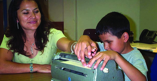 A Hispanic school-age boy takes his mother's hand to show her what he has brailled on his Perkins braillewriter. 