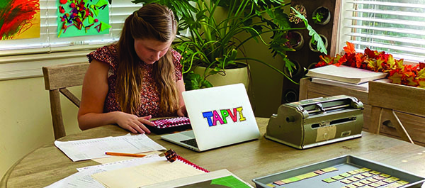 A White teenage girl is at her kitchen table with an abacus during online math instruction. A Perkins braille writer, Wilson Reading System® (WRS) magnetic tiles on a cookie sheet, and other educational materials are on the table. 