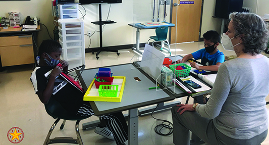 In a classroom, a White teacher wearing a mask sits with two elementary students who are separated by plexiglass and each wearing a mask. Both boys, one African American and one Hispanic, use math manipulatives on APH work trays. Other classmates are on a computer screen mounted to the wall.