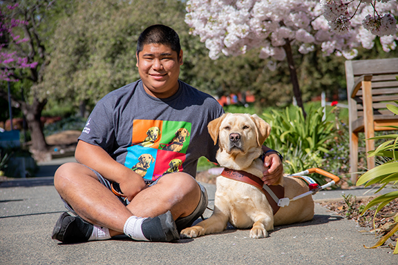 A smiling man sits next to a yellow Lab guide dog.