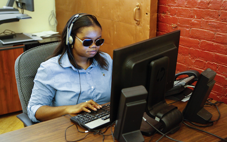 A woman with brown skin wearing dark glasses and headphones sits at a computer.
