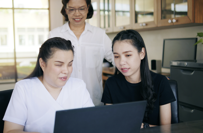Three Asian women co-workers in workplace including a person with blindness using a laptop computer with screen reader program. 