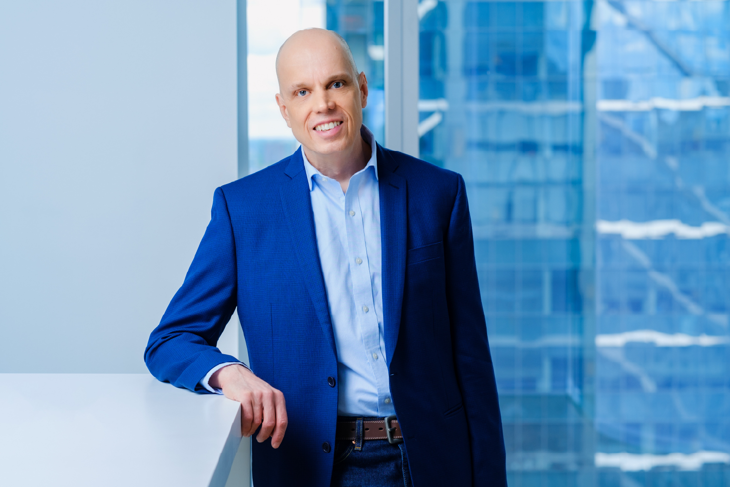 Thomas Wlodkowski in a royal blue suite, smiles while standing in front of a glass window.