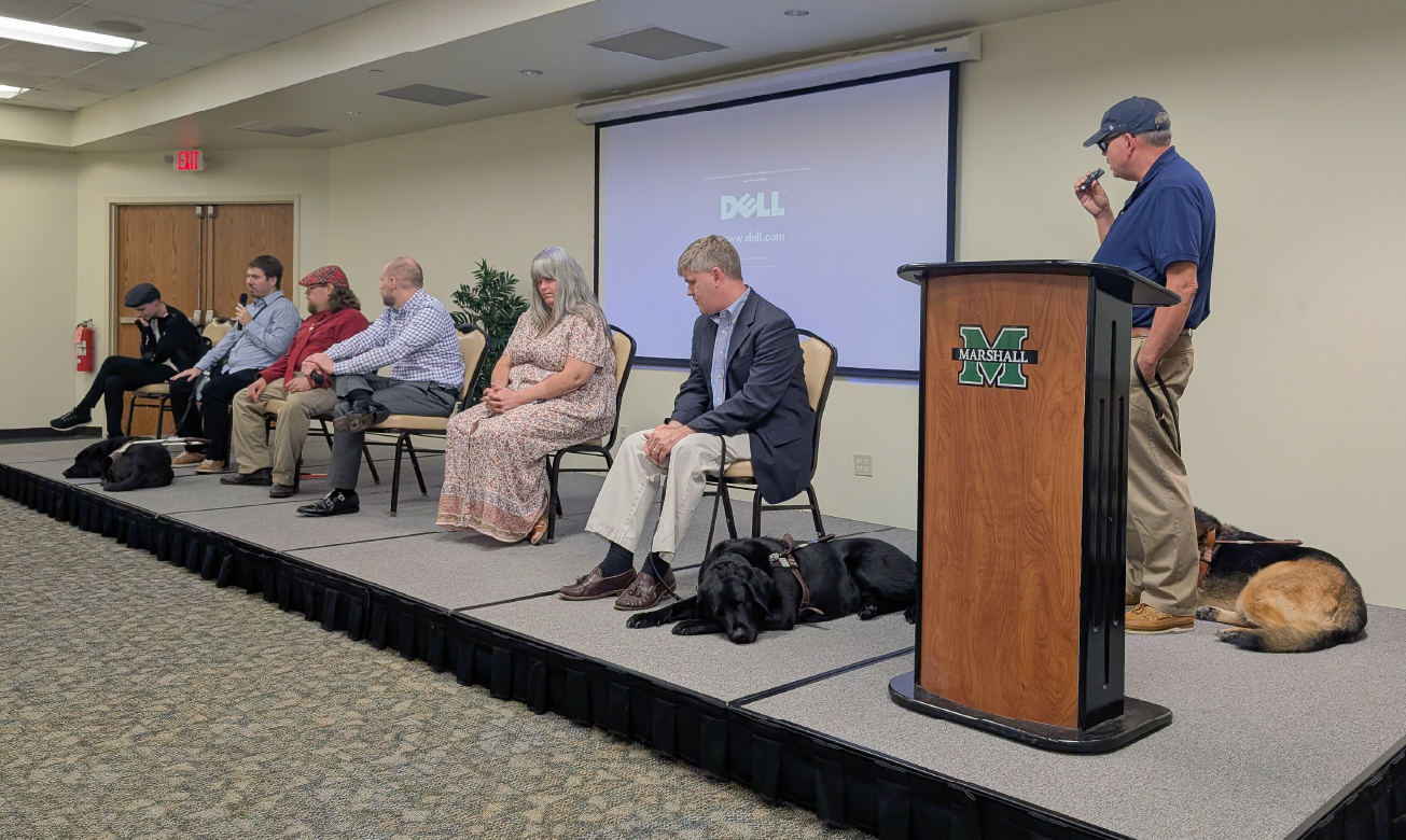 A panelist of 6 individuals sit on a stage, some with their guide dogs. The host stands at a podium, speaking into a mic.