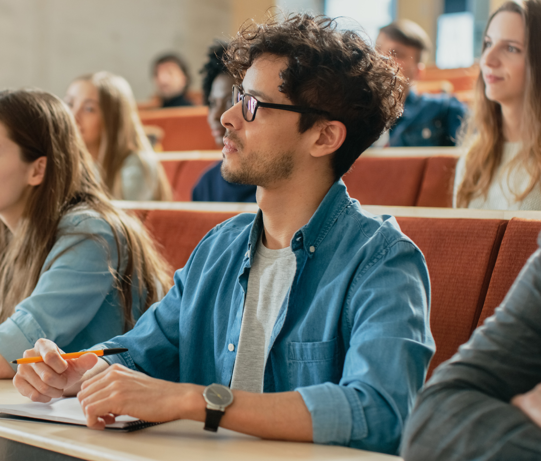 A student wearing glasses sits with other students in a lecture hall.