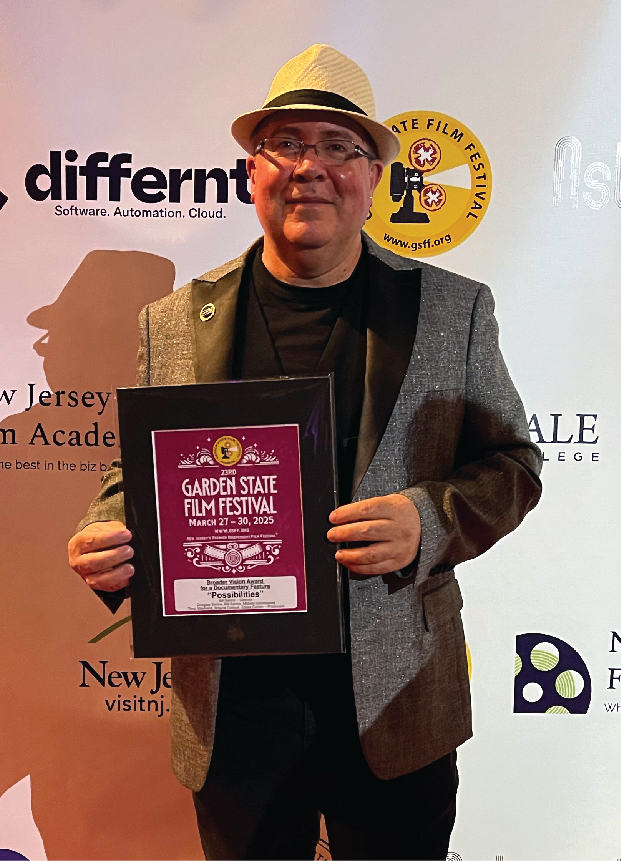 A man with glasses and a hat smiles as he holds an award plaque.