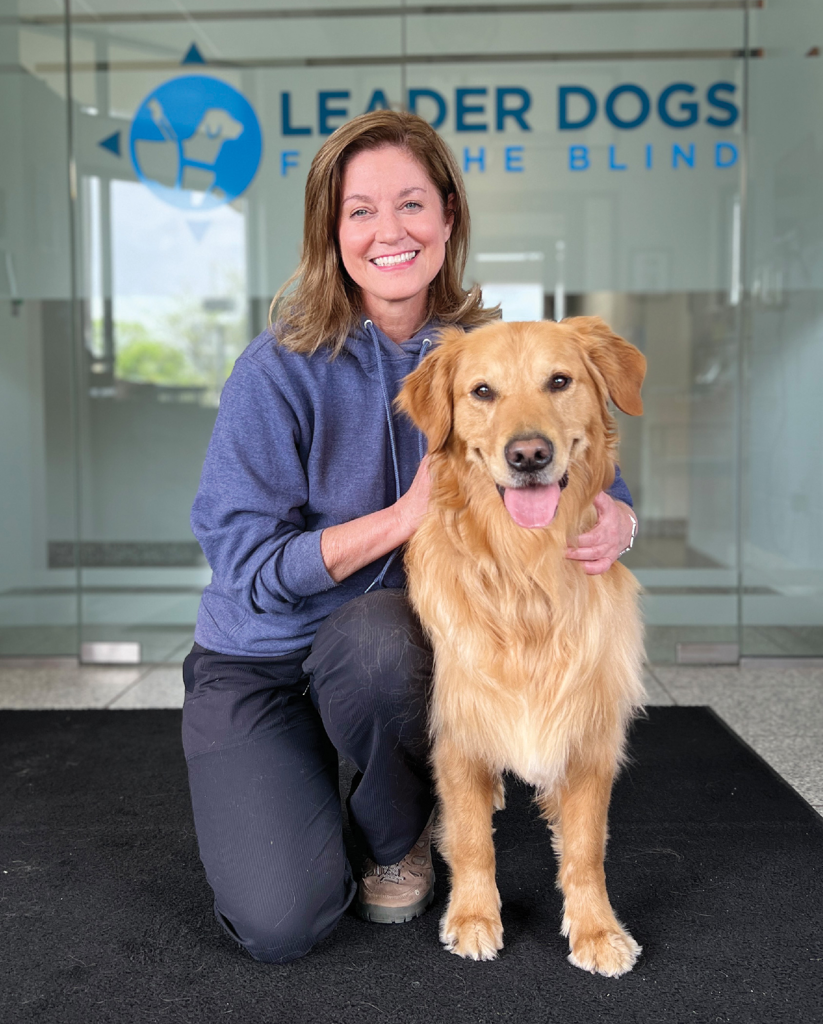 A woman with shoulder length light-brown hair smiles and kneels next to a fluffy golden retriever. A logo in the background reads Leader Dogs for the Blind.