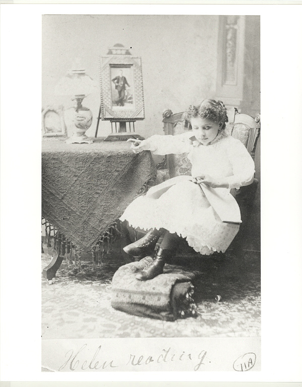Helen Keller as a young girl, seated by a table with a book in her lap. She reads braille text with her left hand and manually signs with her right hand, 1888. Photo courtesy of Perkins School for the Blind