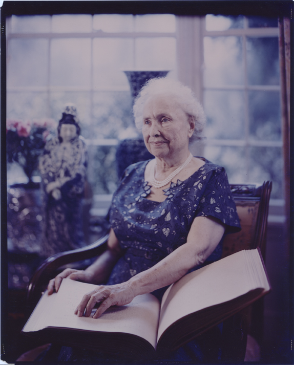 Color photograph of Helen Keller seated in front of a window. She is reading a book in braille, 1960.
