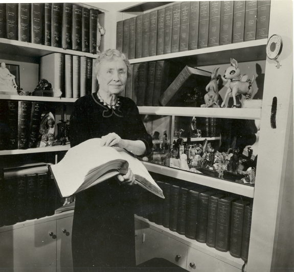 Helen Keller holding a book in braille, while she stands in front of two large bookshelves filled with braille volumes