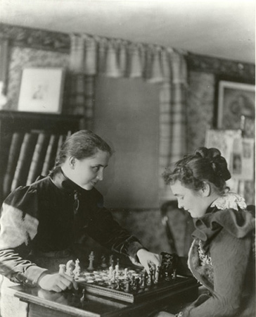 Helen Keller and Anne Sullivan playing chess, 1900. Helen is about to move a white queen. She has already captured one of Anne's black pawns, which is to the right of the board. Helen's hair is pulled into a single braid, down her back. She is wearing a long, dark dress with a high collar and sleeves that puff out from the shoulder to the elbow. Anne's hair is twisted almost like a flower, at the back of her head. She is wearing a dark dress with a white lace collar and dark ruffles around each shoulder.