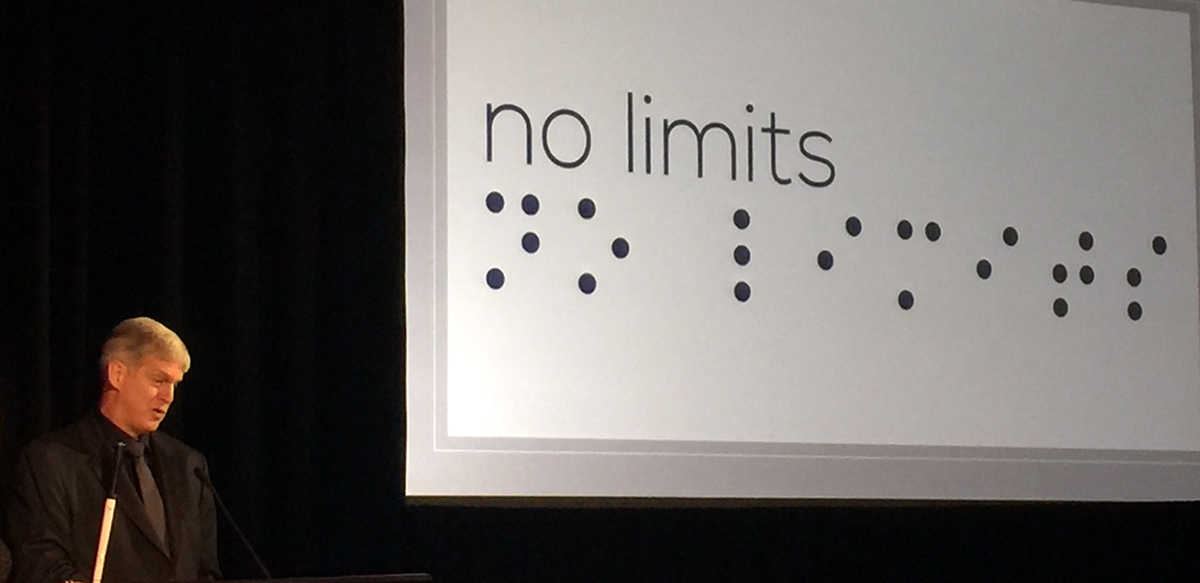 Kirk Adams at the 2017 Helen Keller Achievements Awards with No Limits visible on the screen behind him, in print and simulated braille