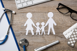 Paper chain cut out of a family of four holding hands. The cut out is on a desk with medical professional, and business tools. 