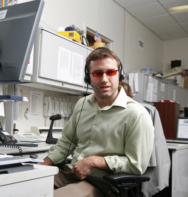 A man wearing tinted glasses and a headset sits in an office.
