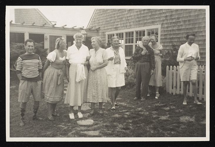 Standing outside Katherine Cornell's home in Martha's Vineyard are left to right: unidentified man (Joseph Lash?), unidentified woman, Eleanor Roosevelt, Helen Keller, Katherine Cornell, unidentified man, Polly Thomson, unidentified woman. They are in the yard, nearby to the wood-sided house. A white picket fence is visible. Everyone is wearing summer clothes and appears relaxed. Keller and an unidentified woman are either side of Roosevelt and all three are linking arms. Polly has her arm around the neck of an unidentified man.