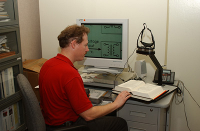 Figure 4: Paul Mogan sitting at a desk with the Jordy mounted on a stand over a large book, while a magnified electrical diagram is displayed on the computer screen.