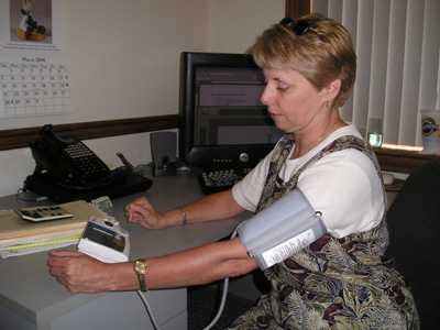 Photo of a woman sitting at a desk with a blood pressure cuff over her upper arm. She is looking at a small rectangular device with a display screen on the desk.