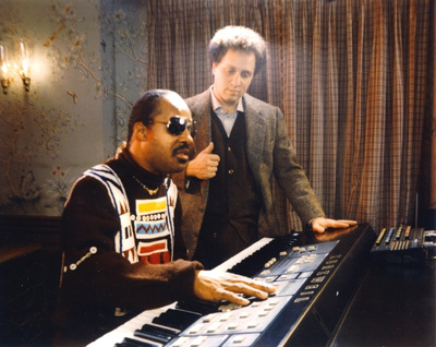 Photo of Stevie Wonder sitting at the keyboard of a large synthesizer with many buttons and switches, while a young Ray Kurzweil looks over his shoulder.