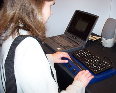 A woman using a braille display with a QWERTY keyboard, attached to a laptop computer.