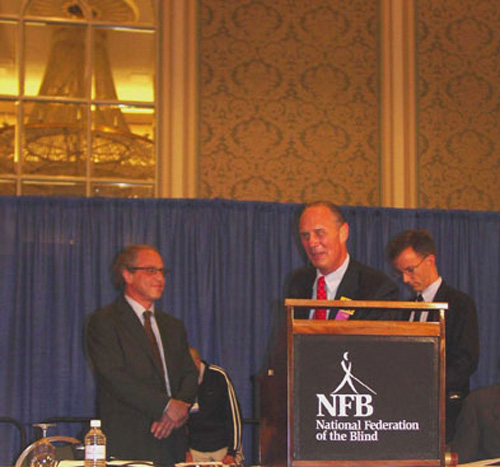 Photo of Jim Gashel at a podium with the NFB logo, standing with Ray Kurzweil.