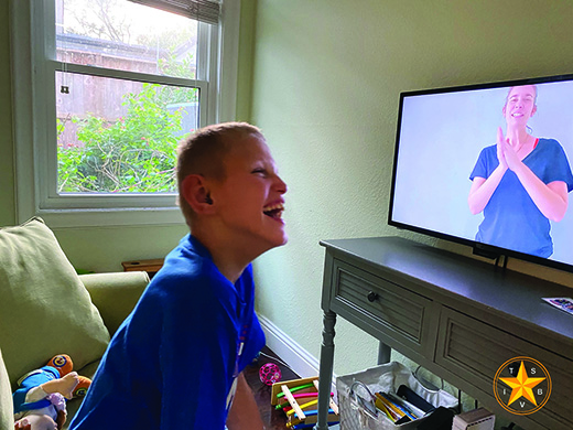 A White deafblind school-age teenager sits in front of a monitor, laughing at the image of his teacher on the screen during a Zoom lesson (courtesy TSBVI).