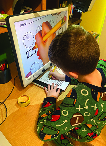 A White school-age boy writes in a box on a workeet about time using a CCTV.