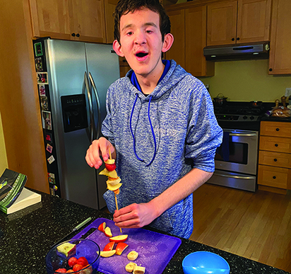 A White school-age teenage boy with additional disabilities in a kitchen puts fruit on skewer.