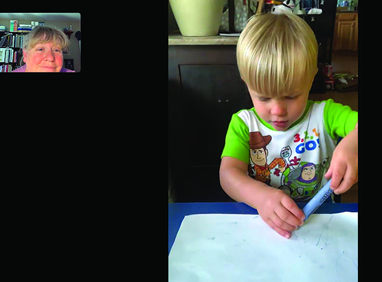 A white toddler colors with a blue crayon as a White TVI watches remotely.” width=“460