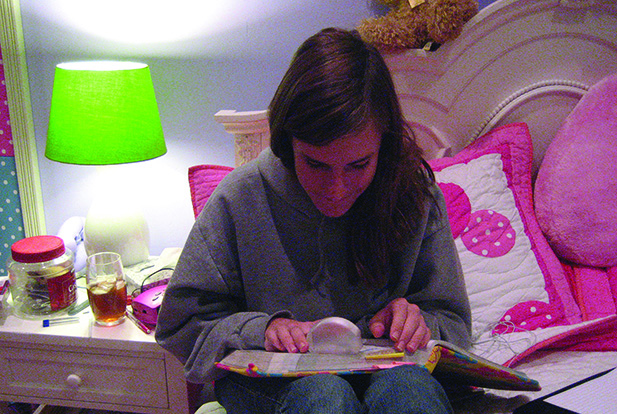 A White school-age teenager sits on a bed reading a book using a dome magnifier.” width=“460