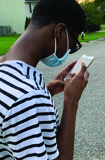 A Black teenage boy wearing a mask is outdoors looking at a smartphone.” width=“460